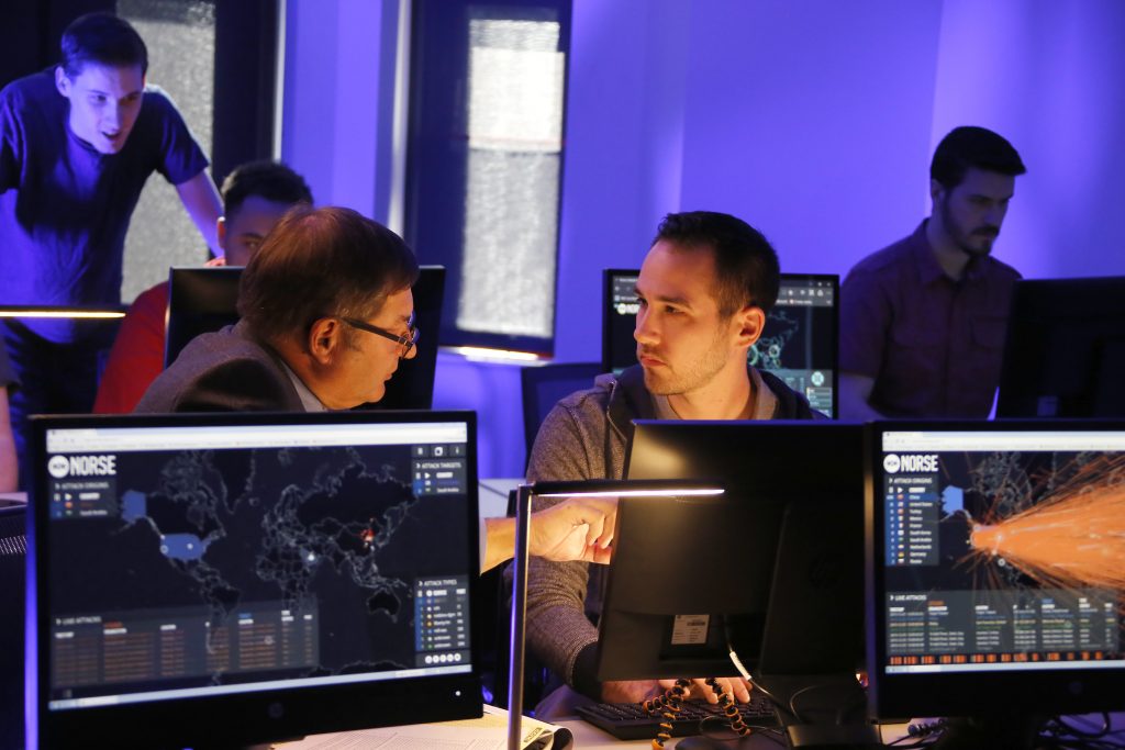 Photo of two men working at a computer in what seems to be a lab or workroom. Other people are in the background.